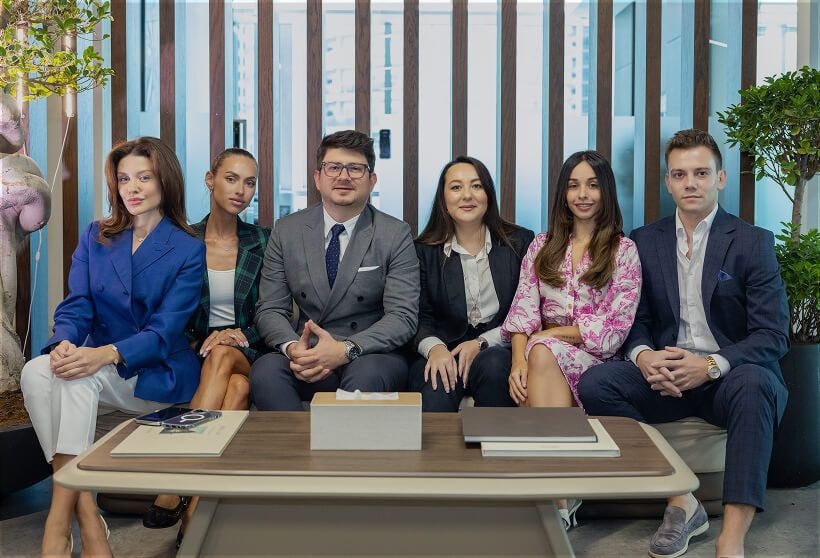A group of six people sits on a modern couch in a professional office setting. They are dressed in formal attire, exuding confidence and teamwork.